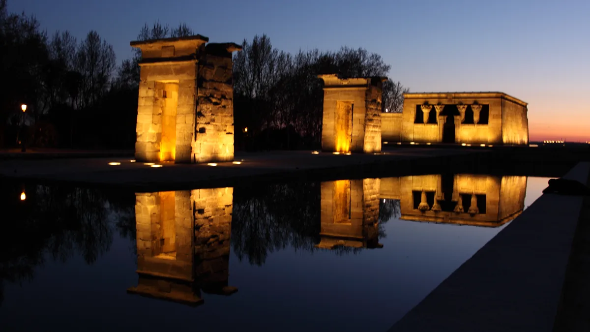 O Templo de Debod ao pôr do sol, iluminado e refletido no espelho d'água, com o céu alaranjado atrás dos pórticos de pedra