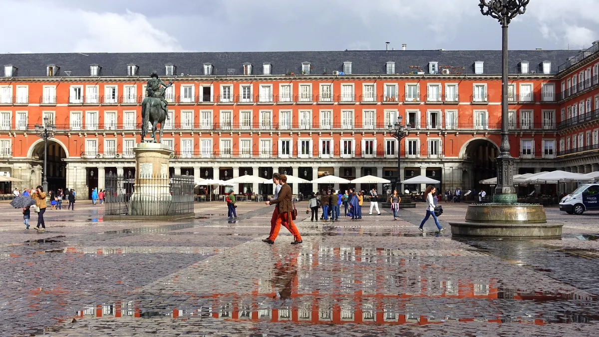 A Plaza Mayor de Madri em dia de chuva, com a fachada vermelha de arcadas refletida no piso molhado, a estátua equestre de Filipe III ao centro e pessoas atravessando
