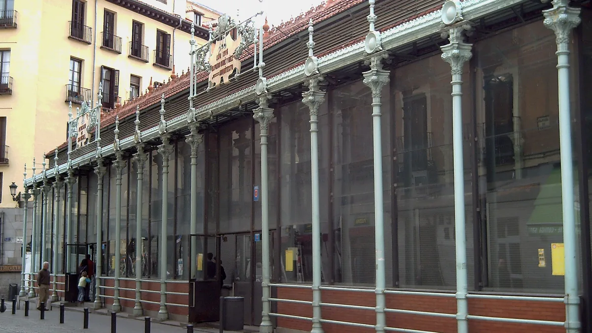 A estrutura de ferro e vidro do Mercado de San Miguel vista de fora, com as colunas pintadas e os toldos sobre a calçada