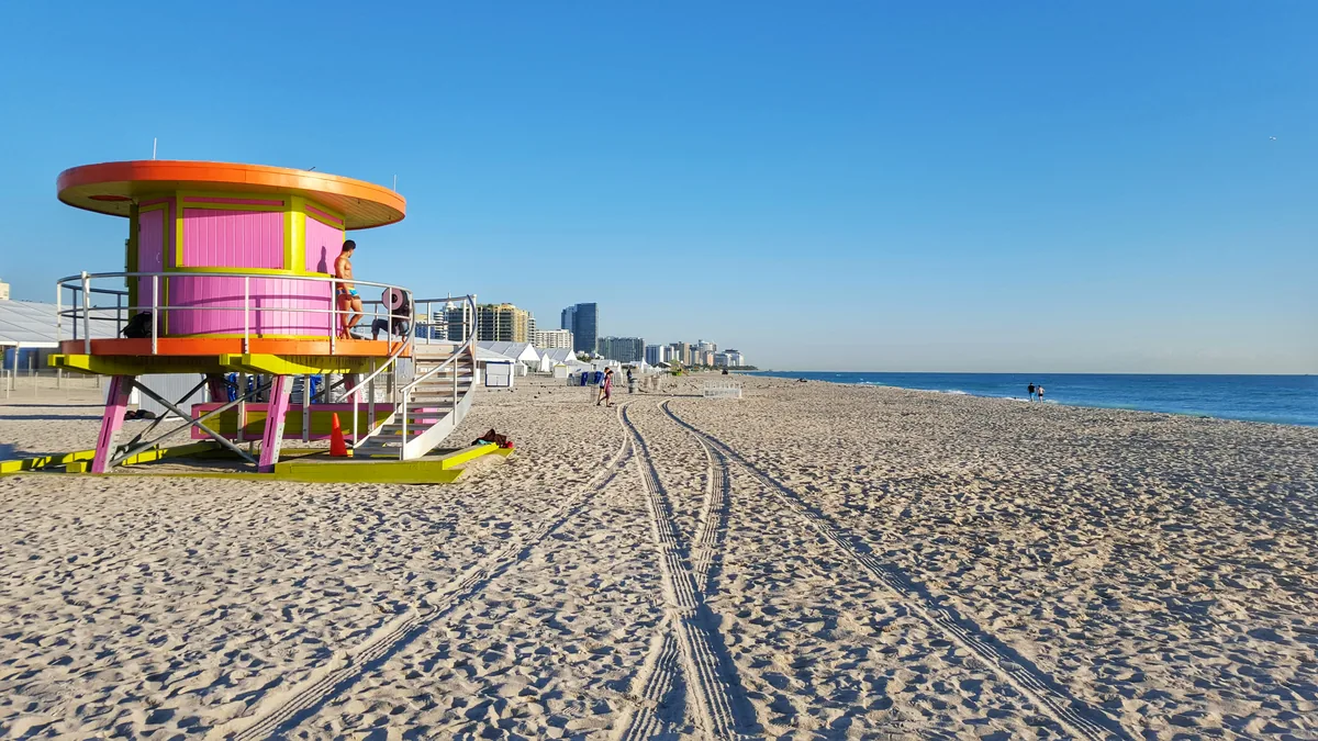 Guarita de salva-vidas pintada em rosa e laranja na areia de South Beach, em Miami, com a praia vazia, rastros de pneu na areia e o skyline de hotéis ao fundo