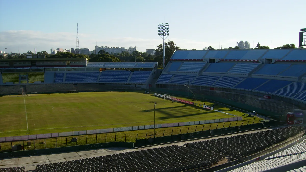 Interior do Estadio Centenario em Montevidéu, com o gramado, a arquibancada azul e o refletor, e a cidade ao fundo