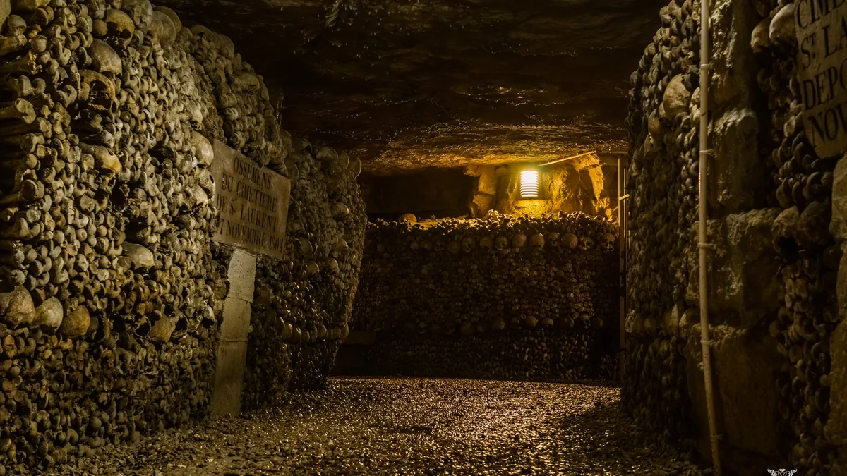 Uma galeria das Catacumbas de Paris, com as paredes de ossos empilhados e a passagem iluminada ao fundo