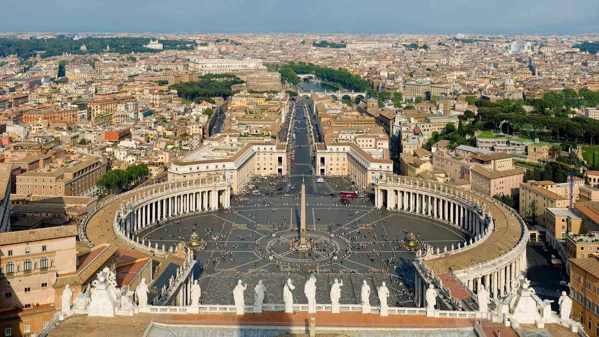 A Praça de São Pedro vista do alto da cúpula da basílica, com a colunata de Bernini em elipse, o obelisco ao centro e a Via della Conciliazione indo até o horizonte de Roma