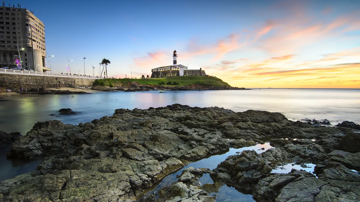 O Farol da Barra ao pôr do sol, visto das pedras da orla, com o céu alaranjado sobre a Baía de Todos os Santos