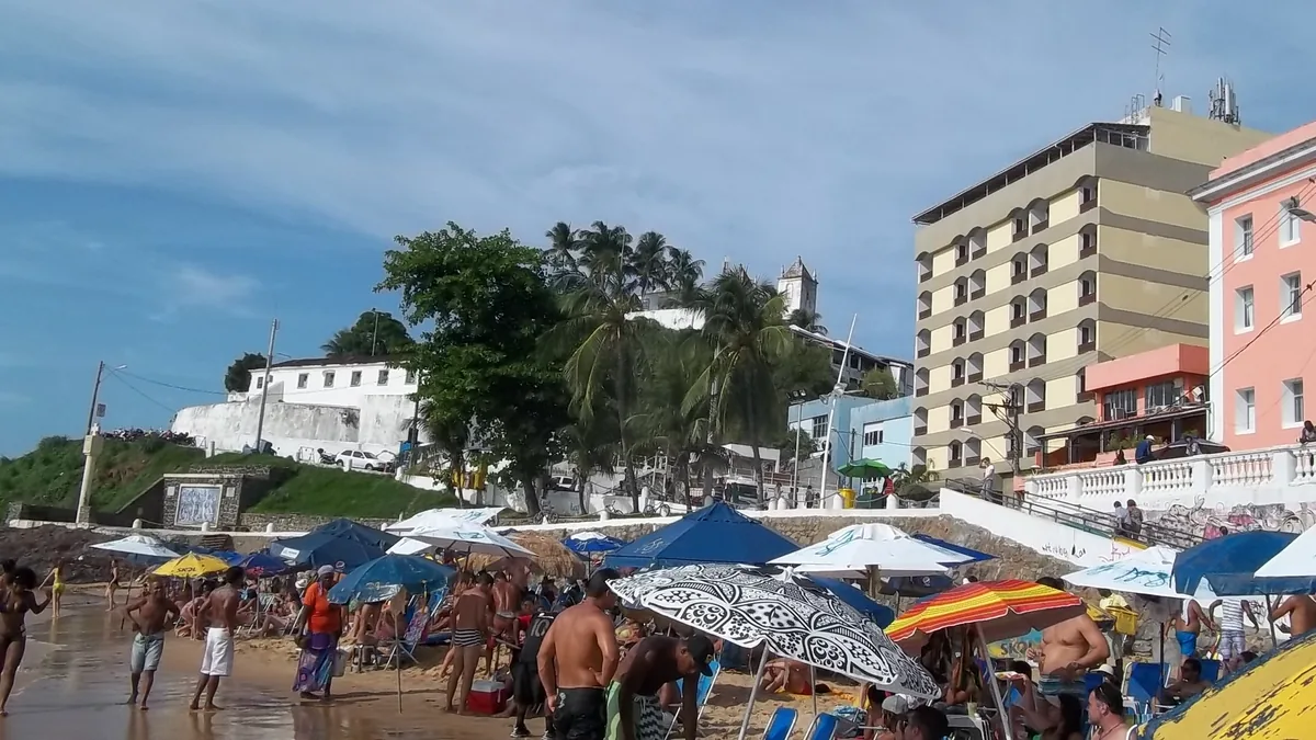A Praia do Porto da Barra cheia, com guarda-sóis coloridos na areia e os prédios da orla ao fundo