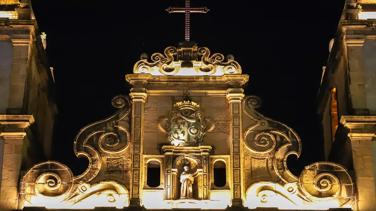 A fachada da Igreja de São Francisco iluminada à noite, com a talha barroca em pedra e a cruz no alto do frontão